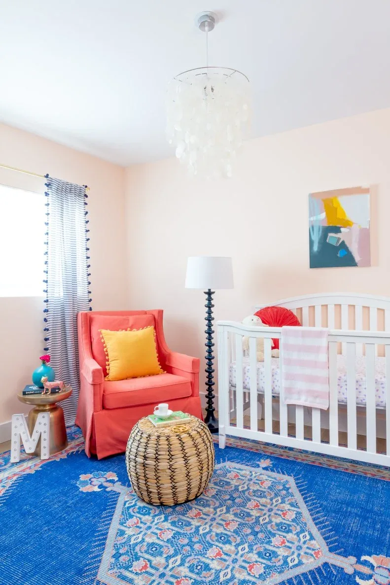 Pale nursery with orange-red adult chair, blue patterned rug, woven round table, white crib and small abstract picture.