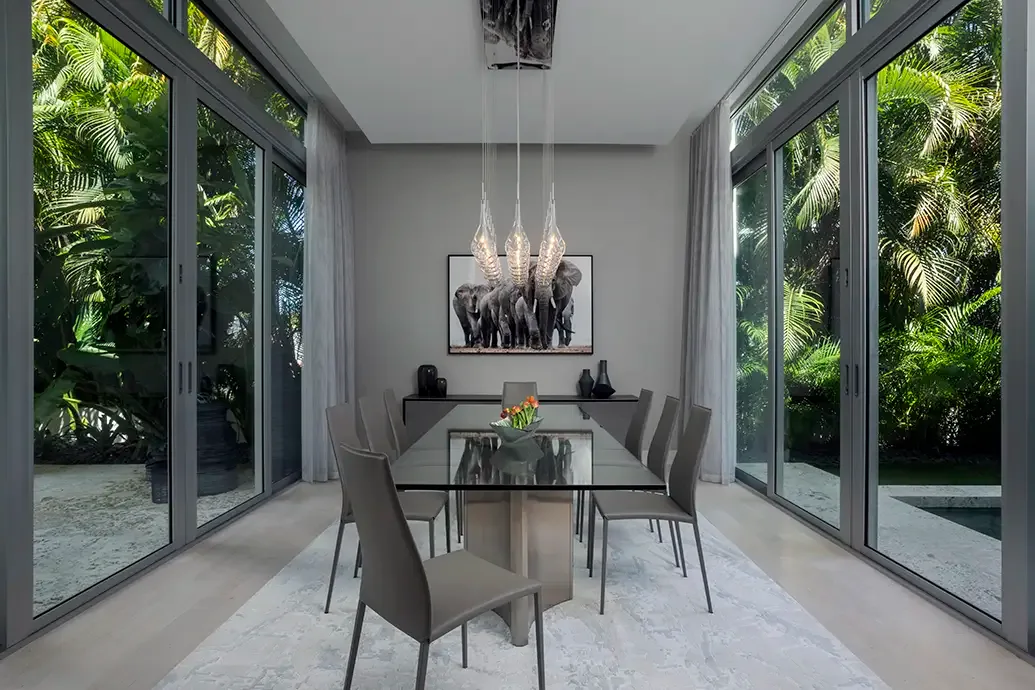 Clear rectangular dining table with gray chairs between full-height windows looking onto greenery
