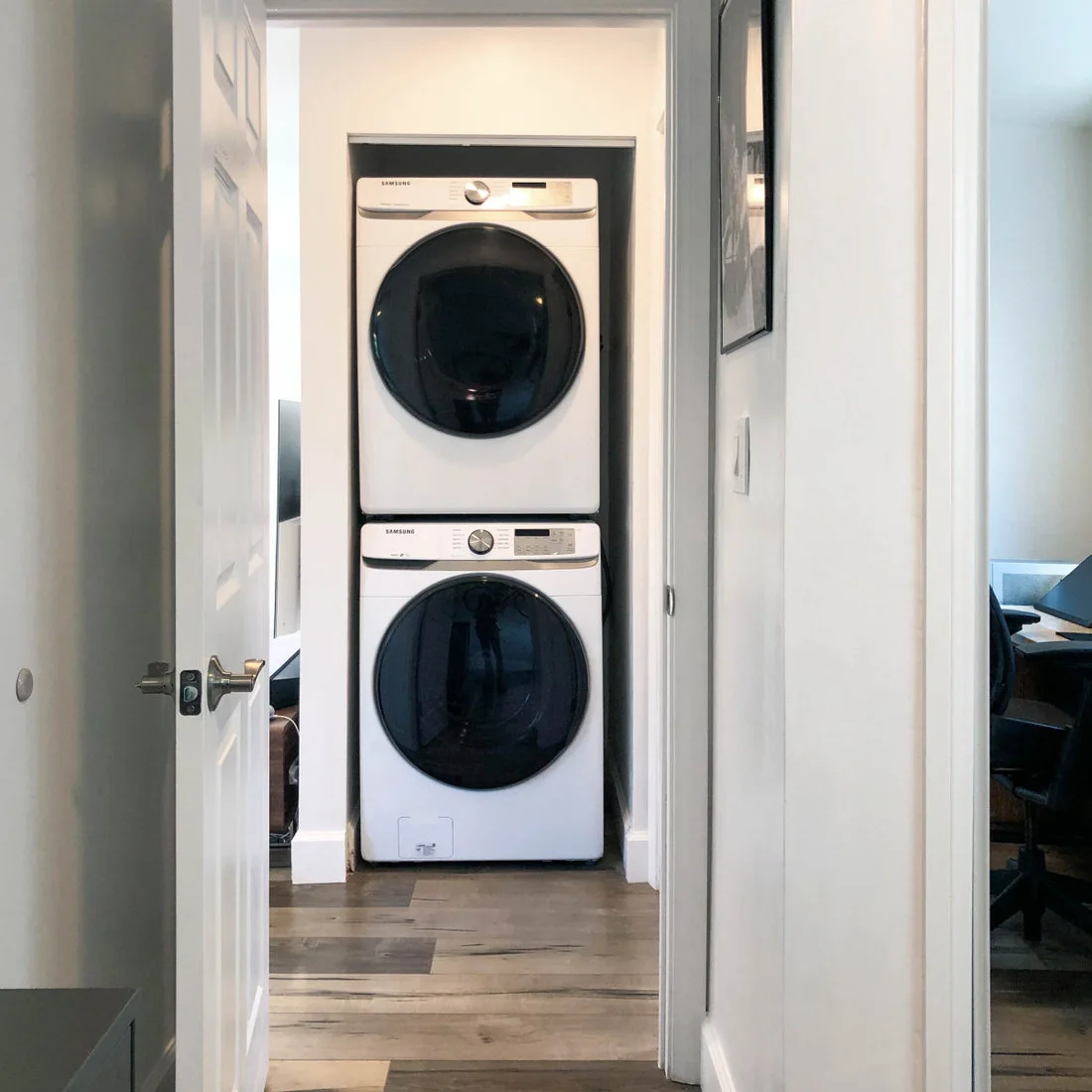 A white washer and dryer are stacked inside a narrow white closet opening onto a pale wood floor.