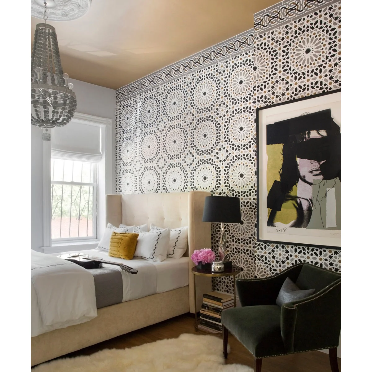 Black and white ornamental wallpaper behind a pale bed and framed portrait with a gold-colored ceiling.