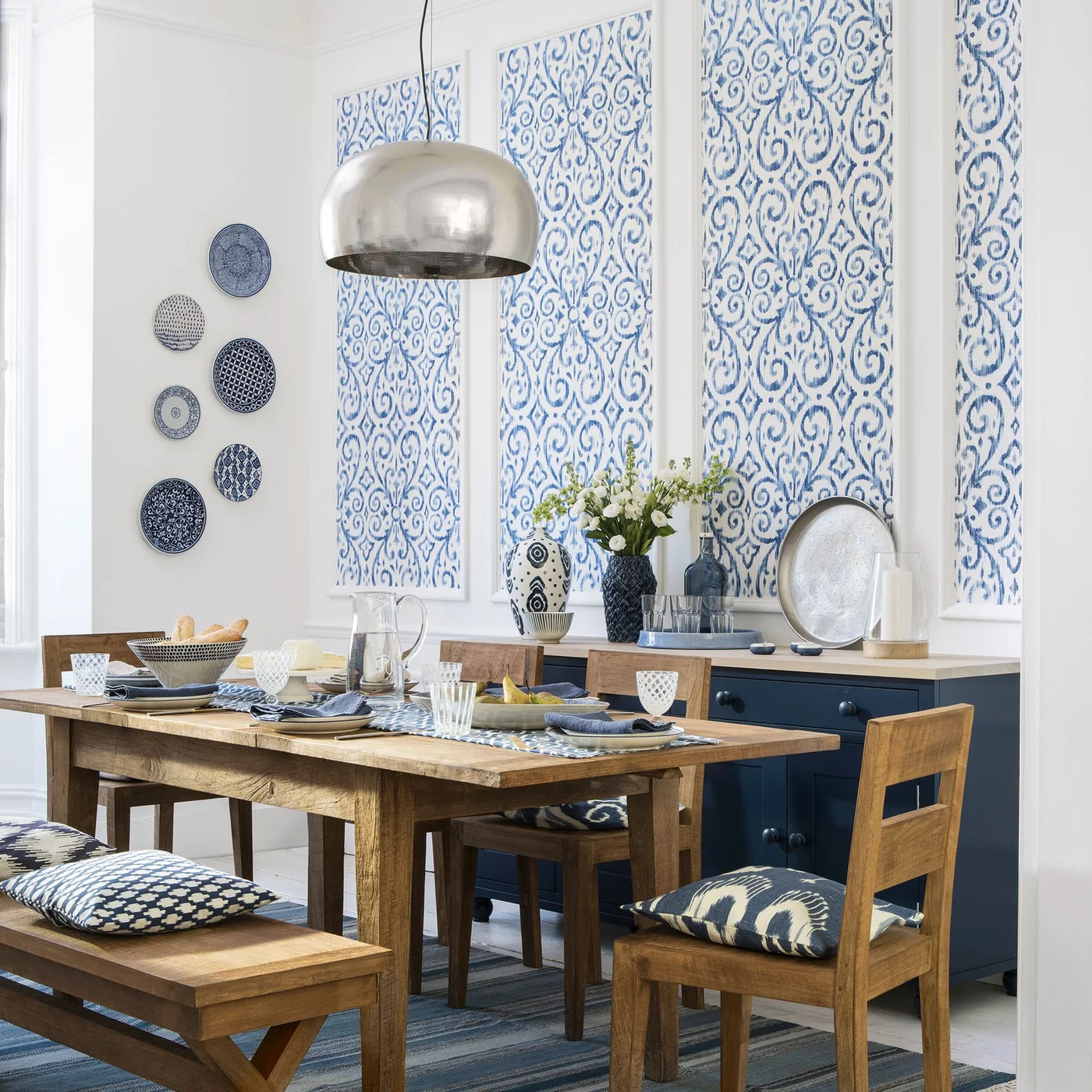 Blue-and-white vessels, glasses on a blue tray and a leaning round platter decorate a navy sideboard beneath blue patterned wall panels.