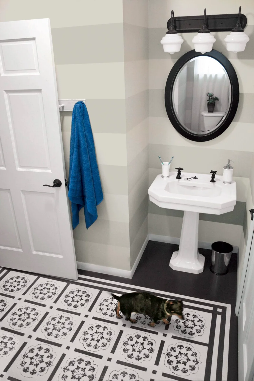 Pale horizontal wall stripes around a white pedestal basin with an oval black mirror and dark three-light fitting.
