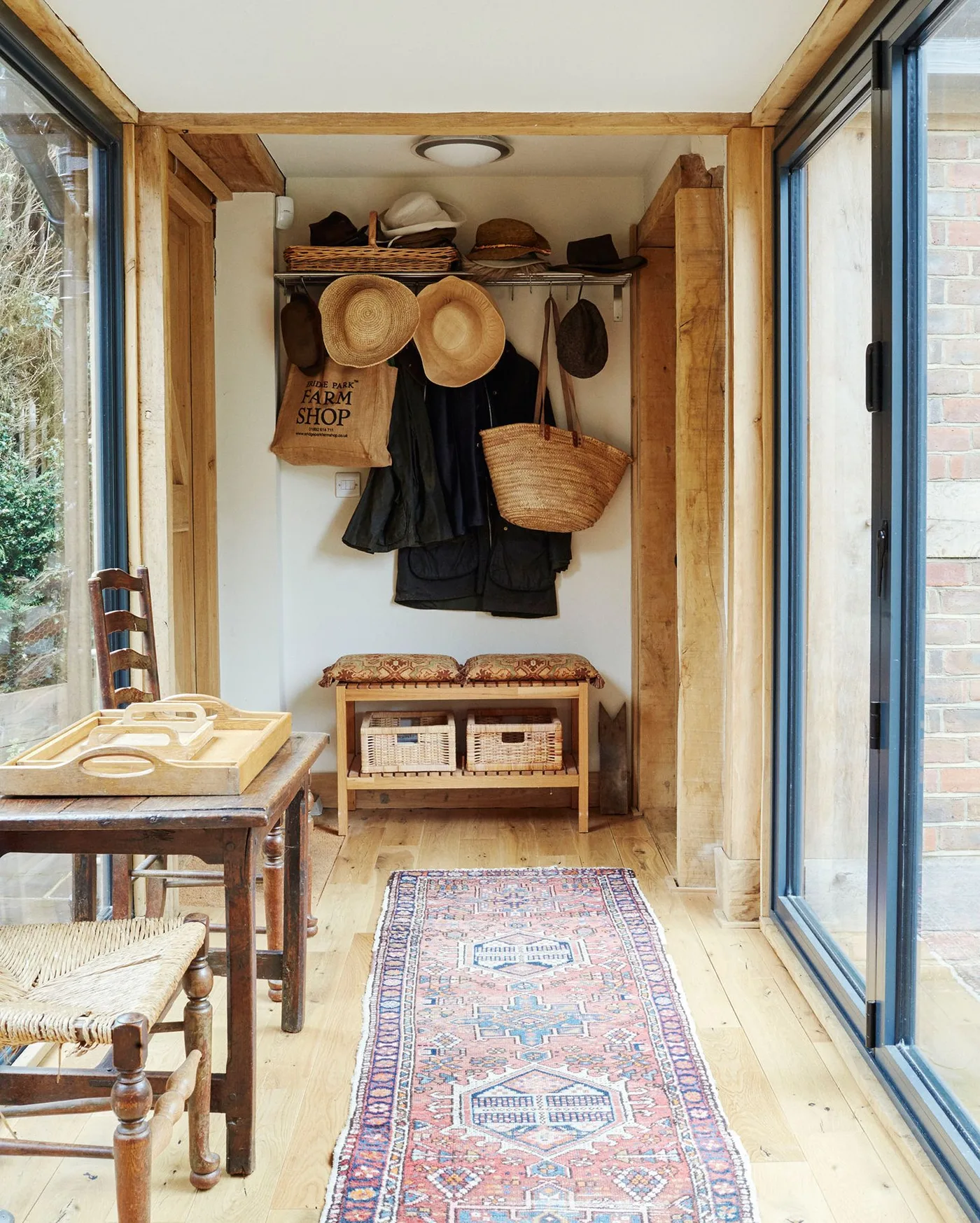 Glazed timber-framed passage with a small cushioned bench, baskets underneath, coats and hats on hooks, wooden table and patterned runner.