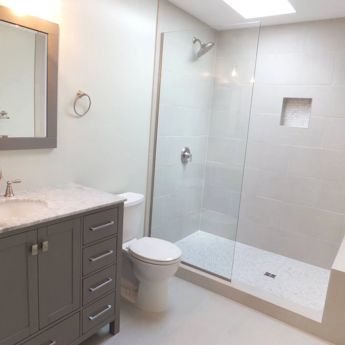 A single clear panel shelters a white tiled shower tray beside a gray vanity and toilet, with an open entrance on the right.