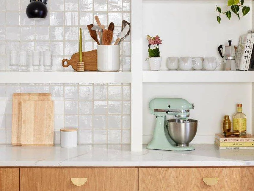 Glossy white square tiles form a backsplash behind open shelves beside timber drawers and a pale green mixer.