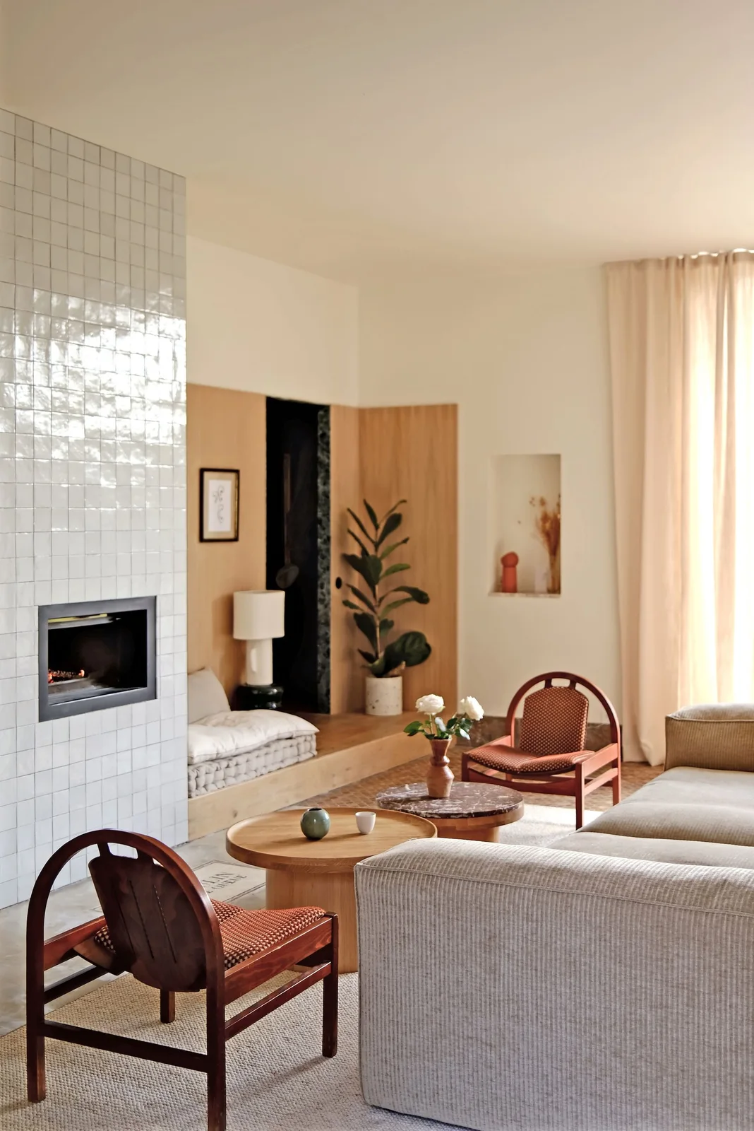 Small pale glossy square tiles cover a fireplace wall beside a built-in wood bench, a light sofa, reddish wood chairs and round tables.