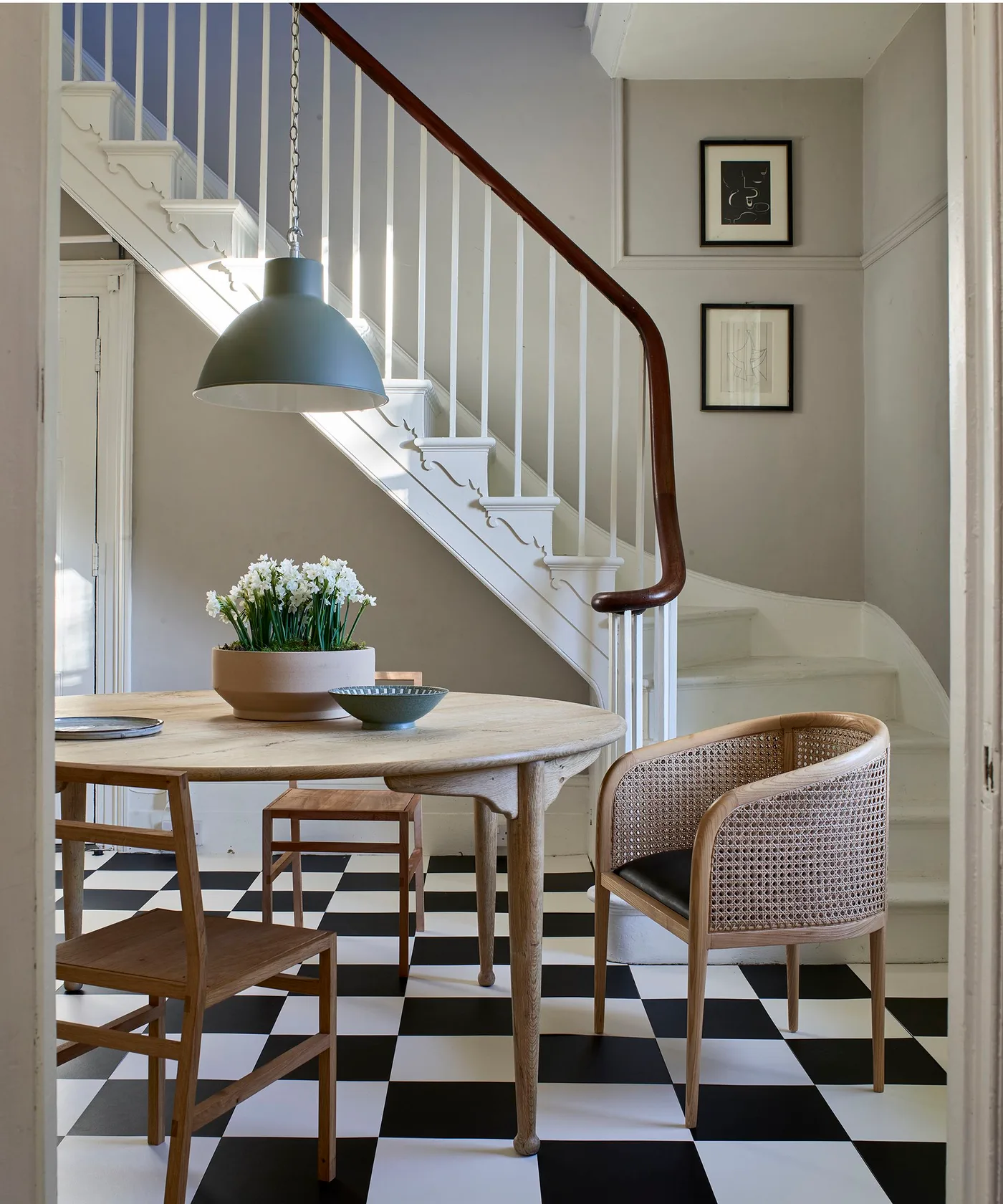 Country dining corner beneath a staircase with black-and-white checked flooring, round pale wooden table, mixed wooden chairs and a green pendant light.