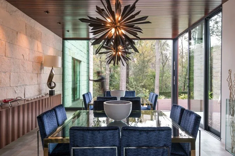 Rectangular glass dining table with deep blue chairs under starburst lights beside full-height windows and a textured wall