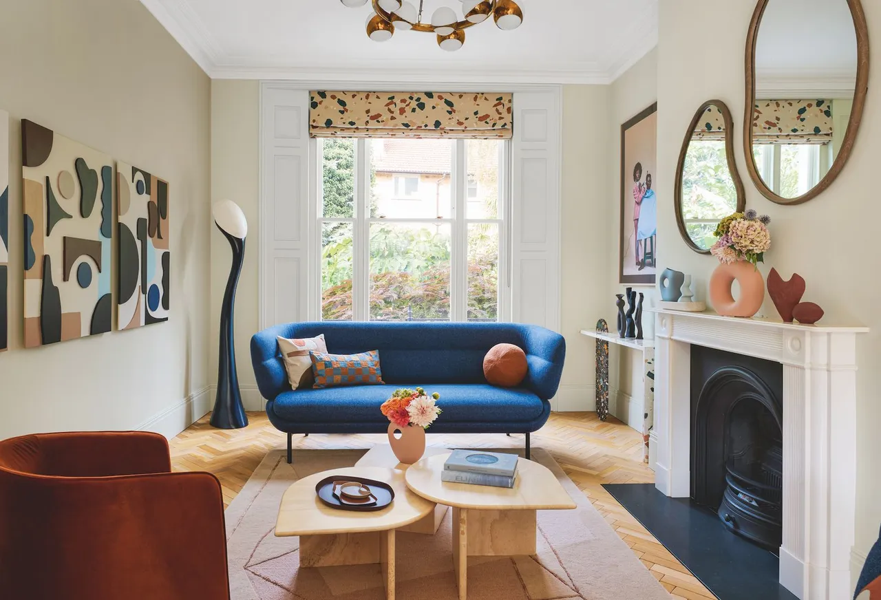 A small blue sofa stands beside a pale fireplace with warm-colored cushions and simple tables.