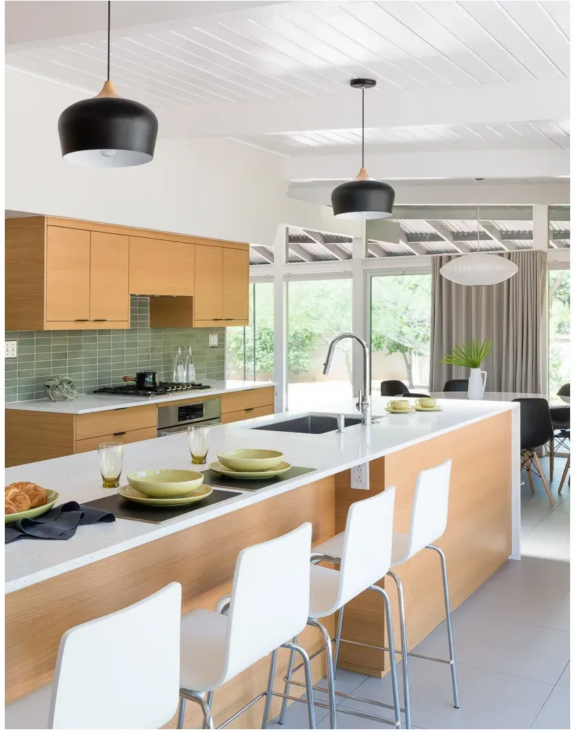 Pale wood cabinets and a white-topped island with white stools beneath black pendants and a muted green backsplash.