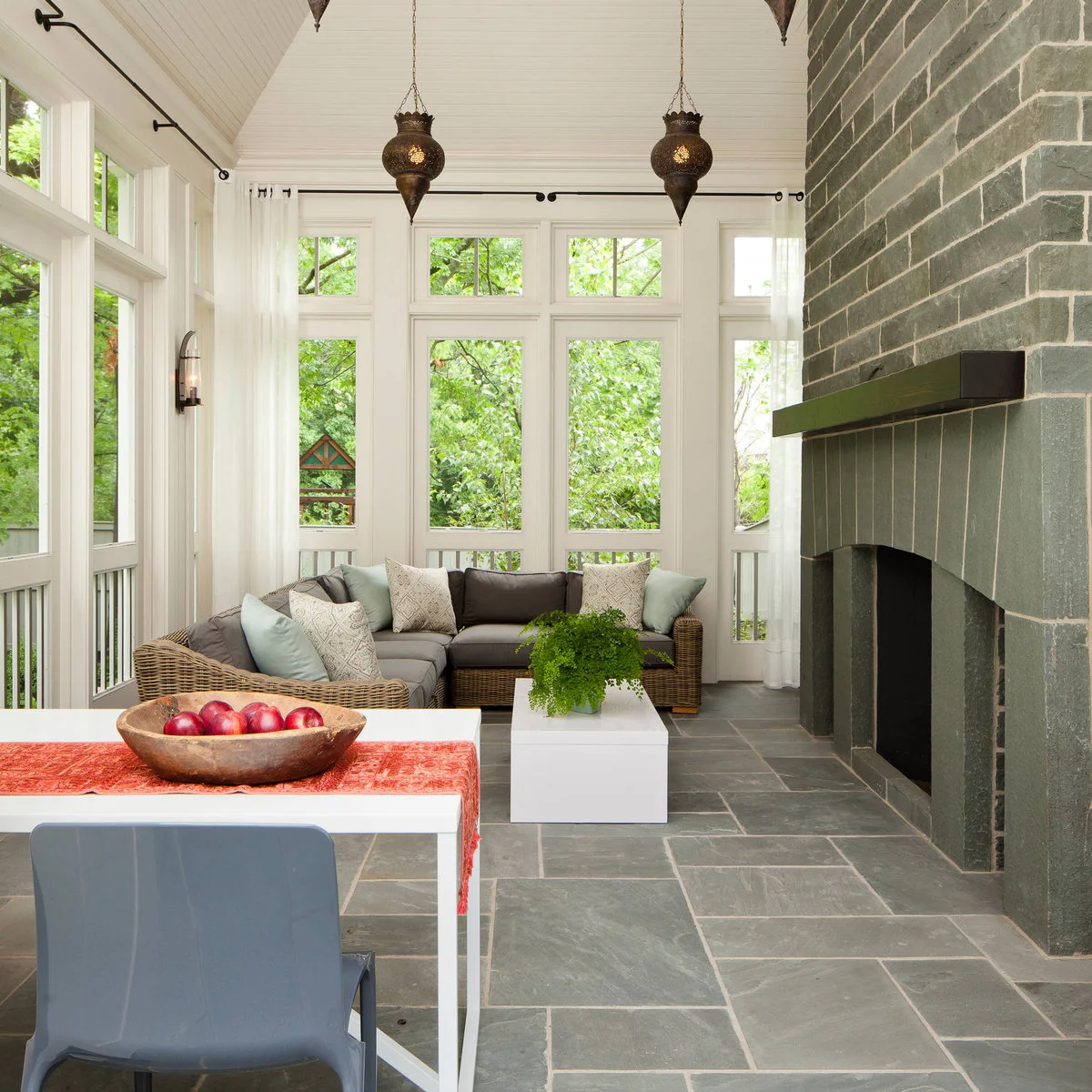 A wicker sectional and low white table beside a dark stone fireplace, with a separate dining surface in the foreground.
