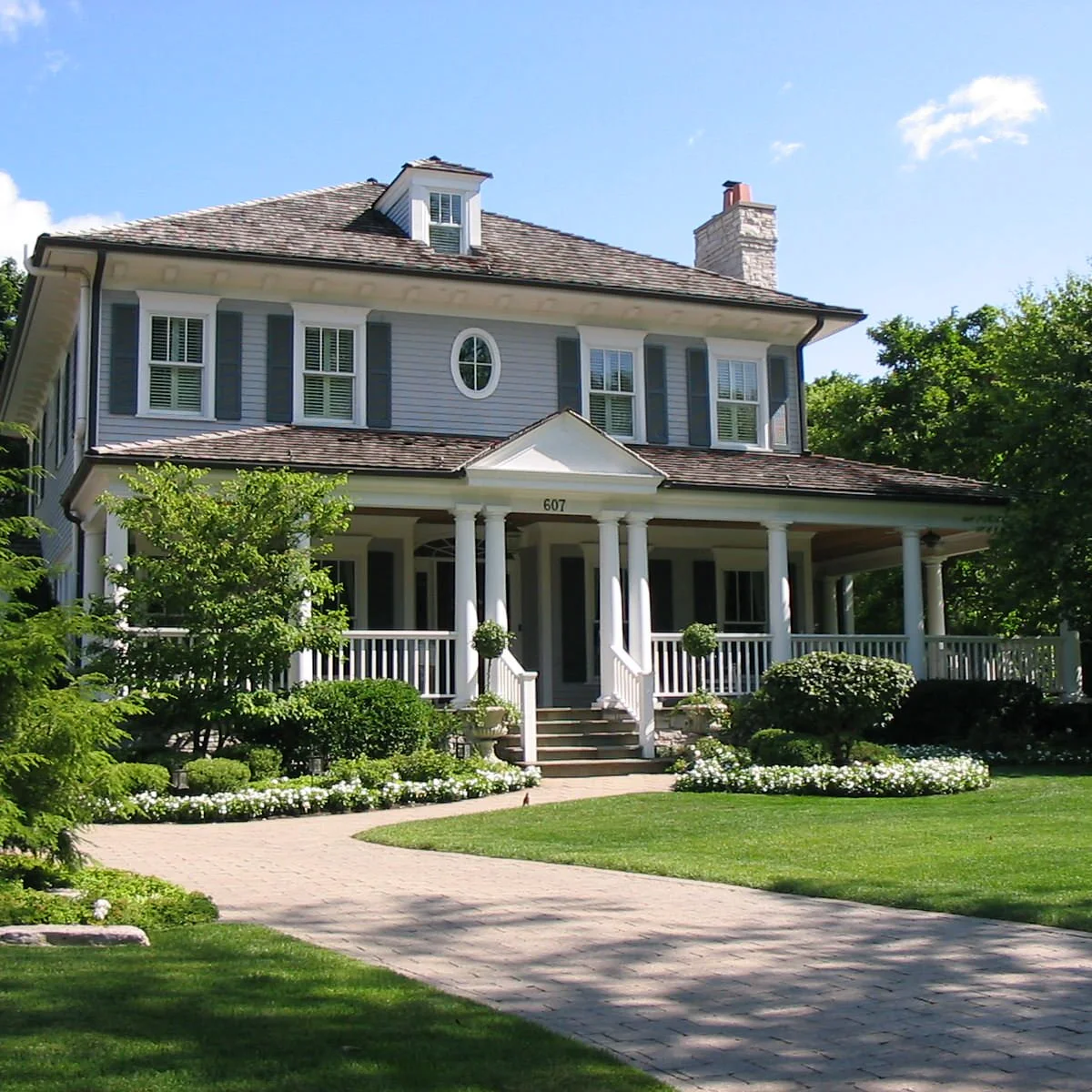 A gray house with a wraparound white porch, rounded mature shrubs and a low white-flowered border beside the front walk.
