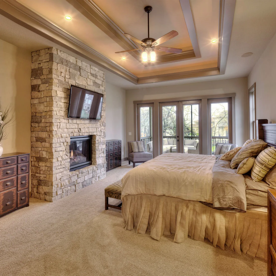 A neutral bedroom with a stone fireplace, timber bed and two small chairs beside glazed doors