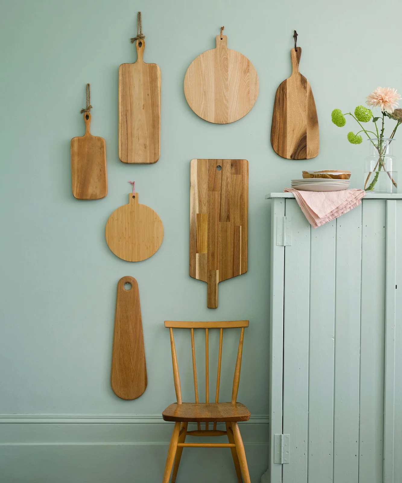 Wooden serving boards in varied shapes hung on a pale green wall beside a matching cabinet and wooden chair.