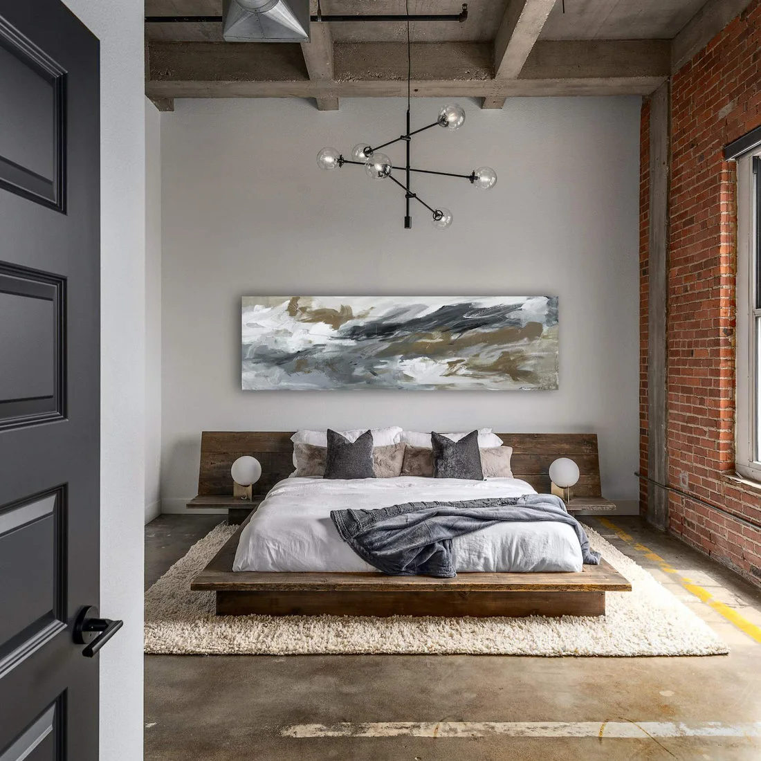 A low wooden bed with white bedding sits beneath a rough concrete ceiling beside exposed brick and a branching black pendant