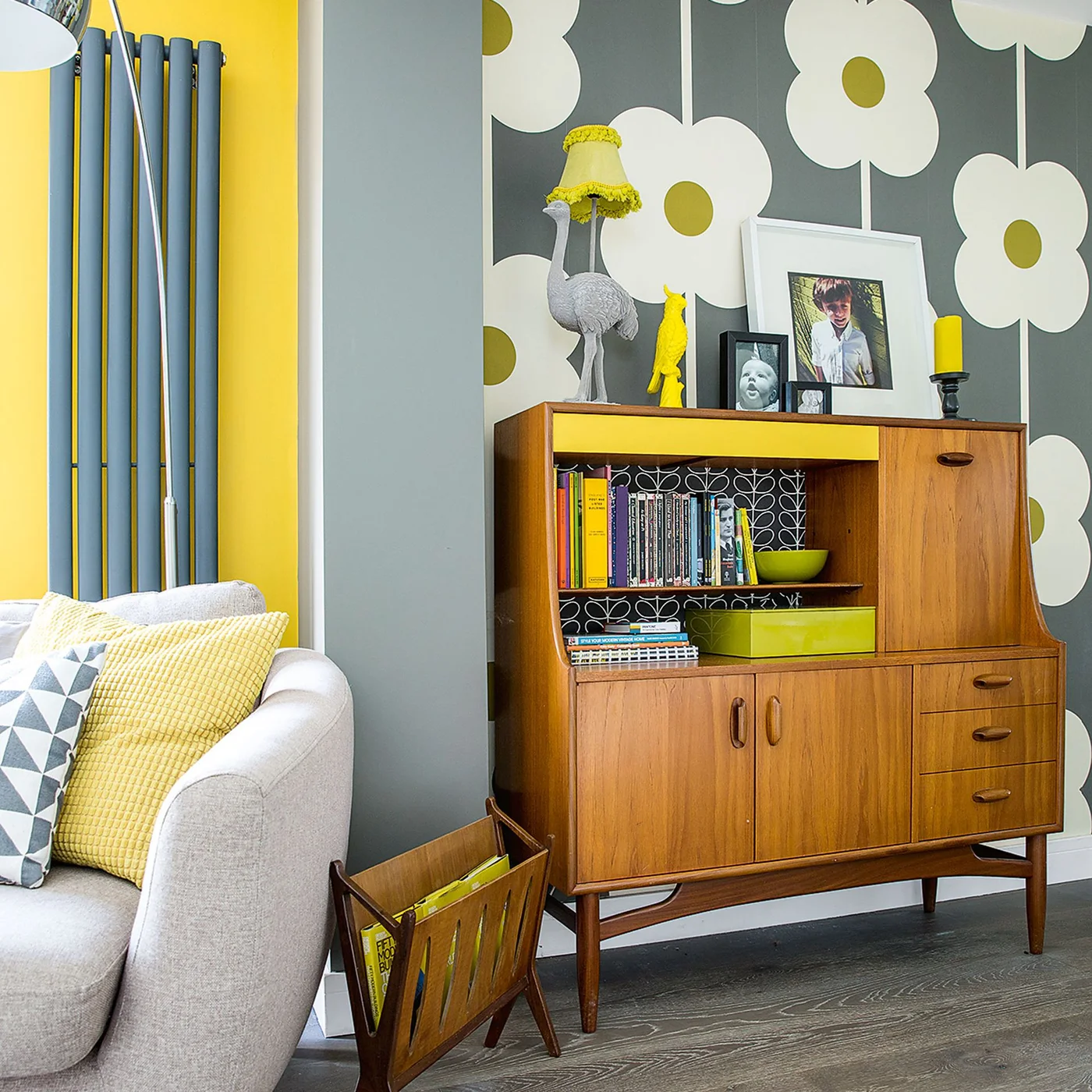 Oversized white flowers on a gray wallpaper sit behind a wooden cabinet beside a yellow wall and pale sofa