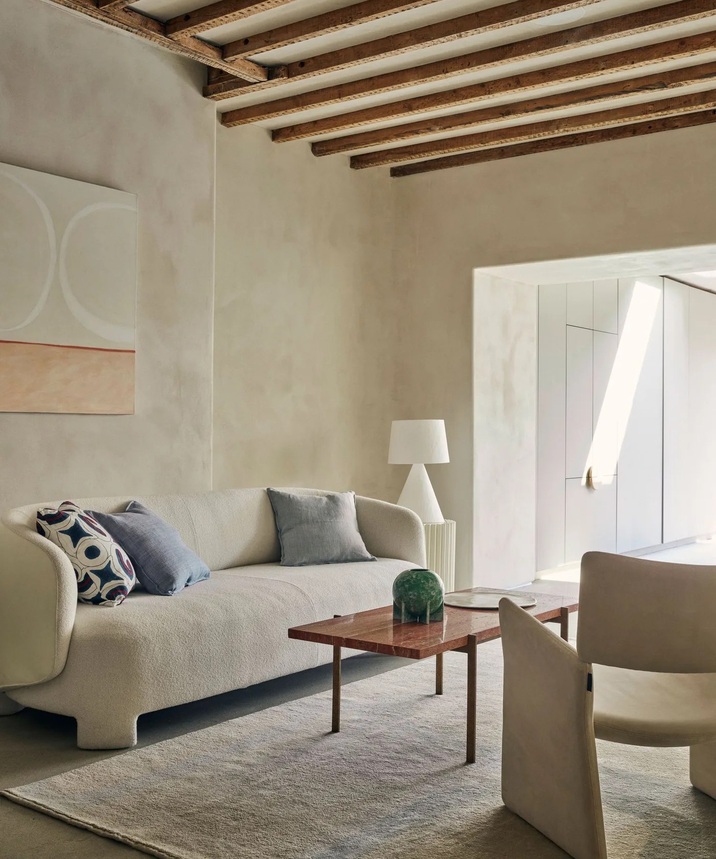 Warm exposed wood ceiling beams over a pale curved sofa, muted rug and reddish rectangular coffee table