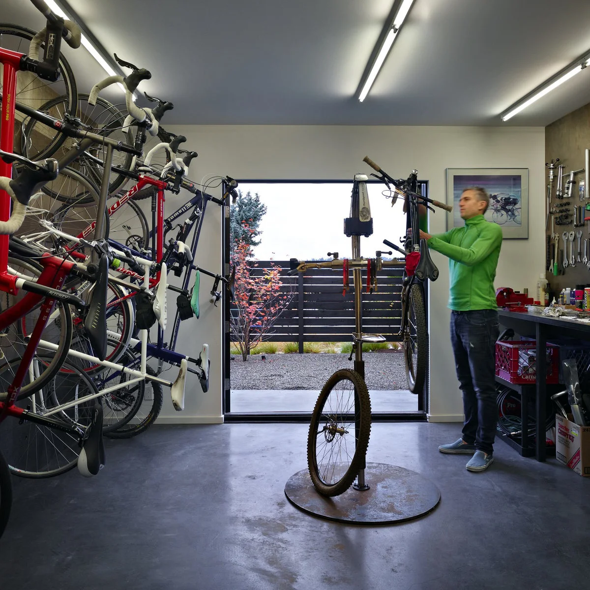 Narrow bicycle workshop with vertical bike storage on one wall, a tool bench opposite and an open exterior doorway.