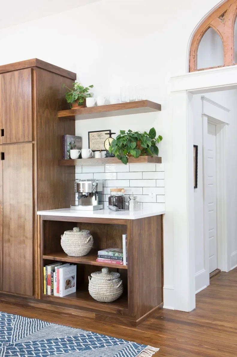 Two wood-toned floating shelves with cups and plants above a coffee counter beside a tall wooden cupboard