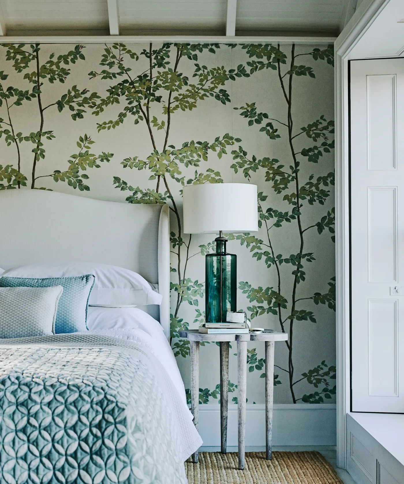 Green tree-pattern wallpaper behind a pale headboard, white bedding and a tall green glass lamp.