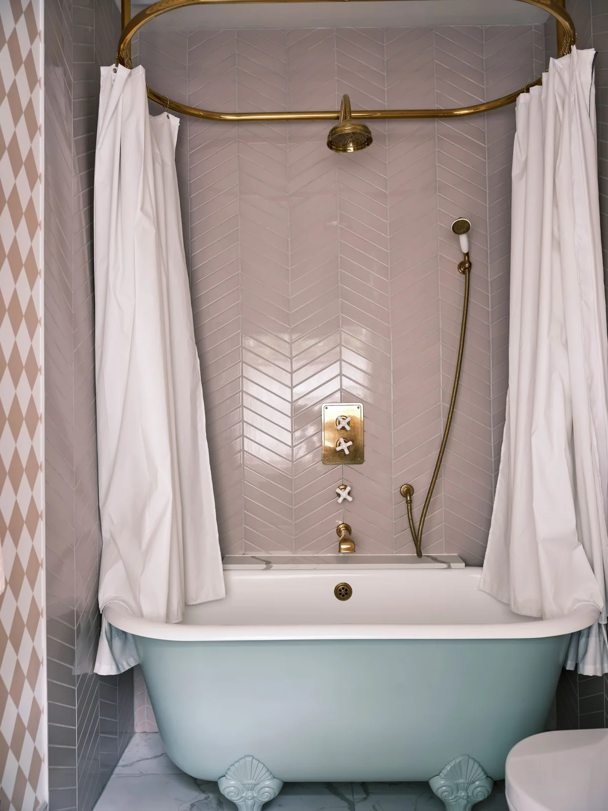 Pale pink herringbone tiles surround a bath below a gold-colored oval shower rail and white curtains