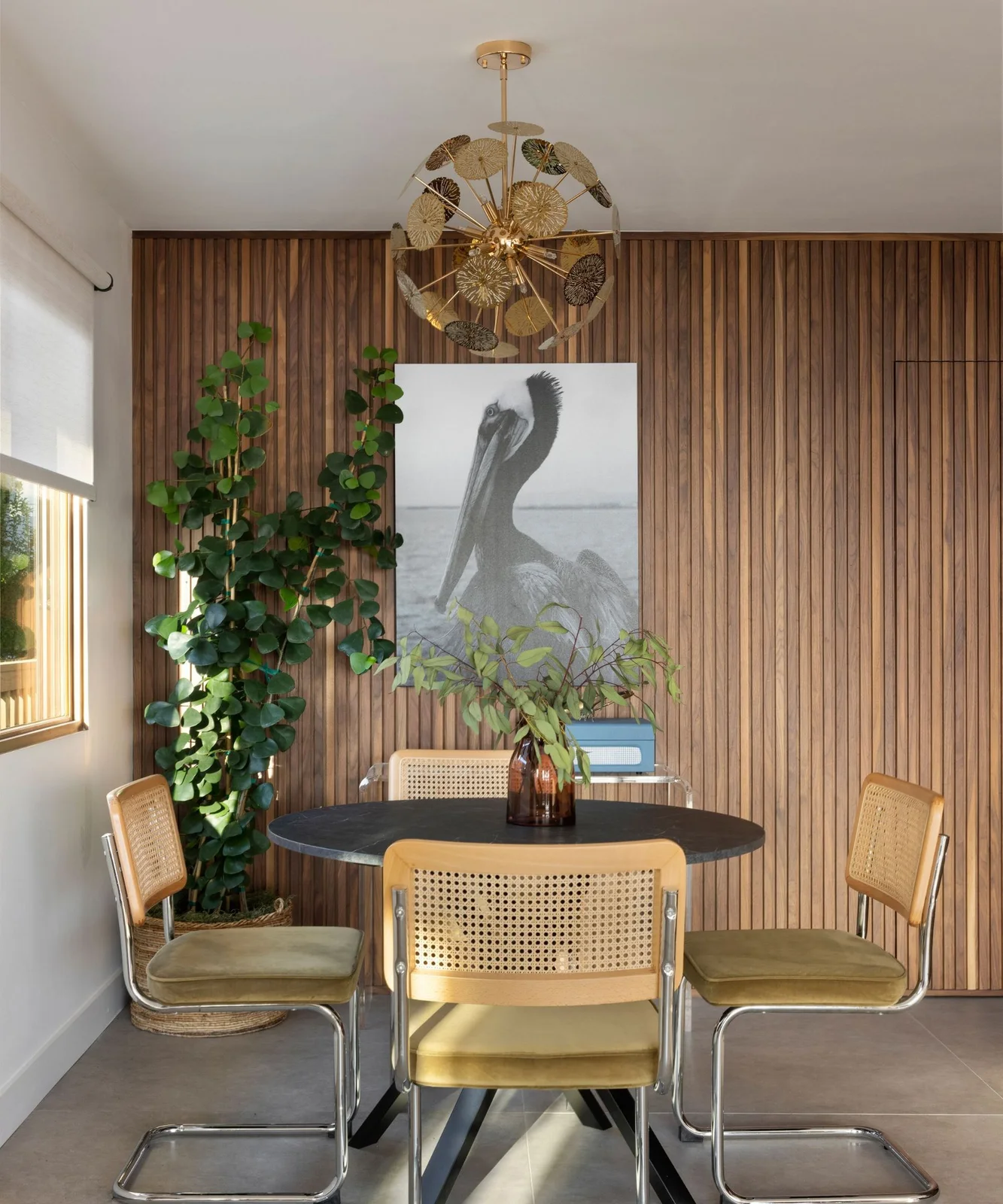 A dark round dining table stands among woven-back chairs with tubular metal frames beside vertical wood panels, a bird print and decorative warm-colored chandelier