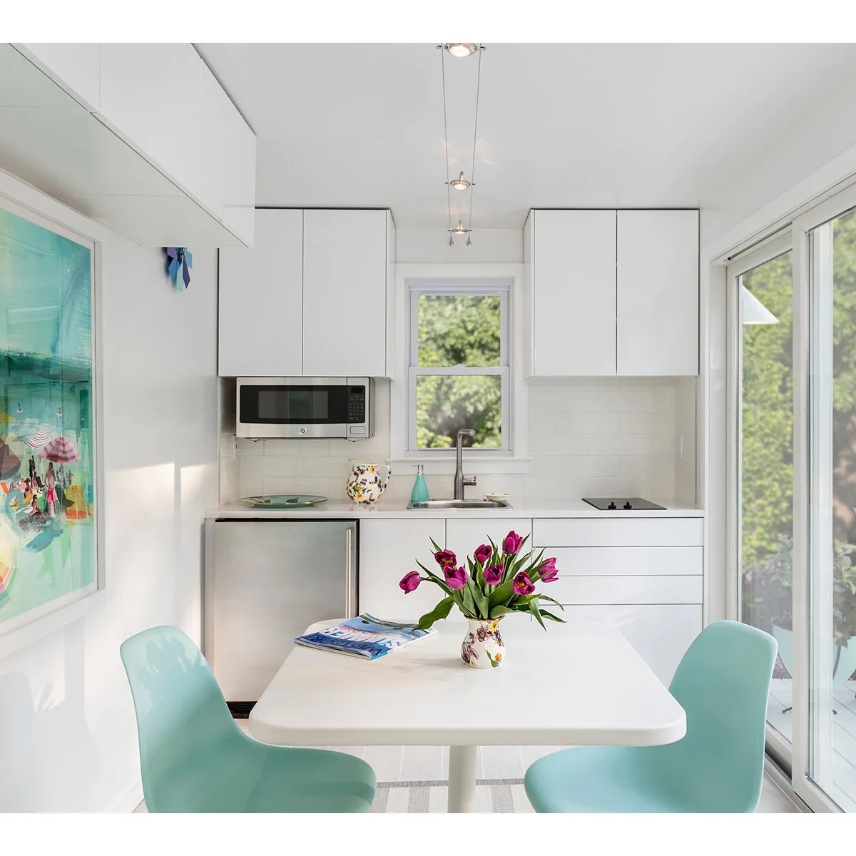 White kitchenette with a small cooktop, sink and refrigerator behind a table with aqua chairs