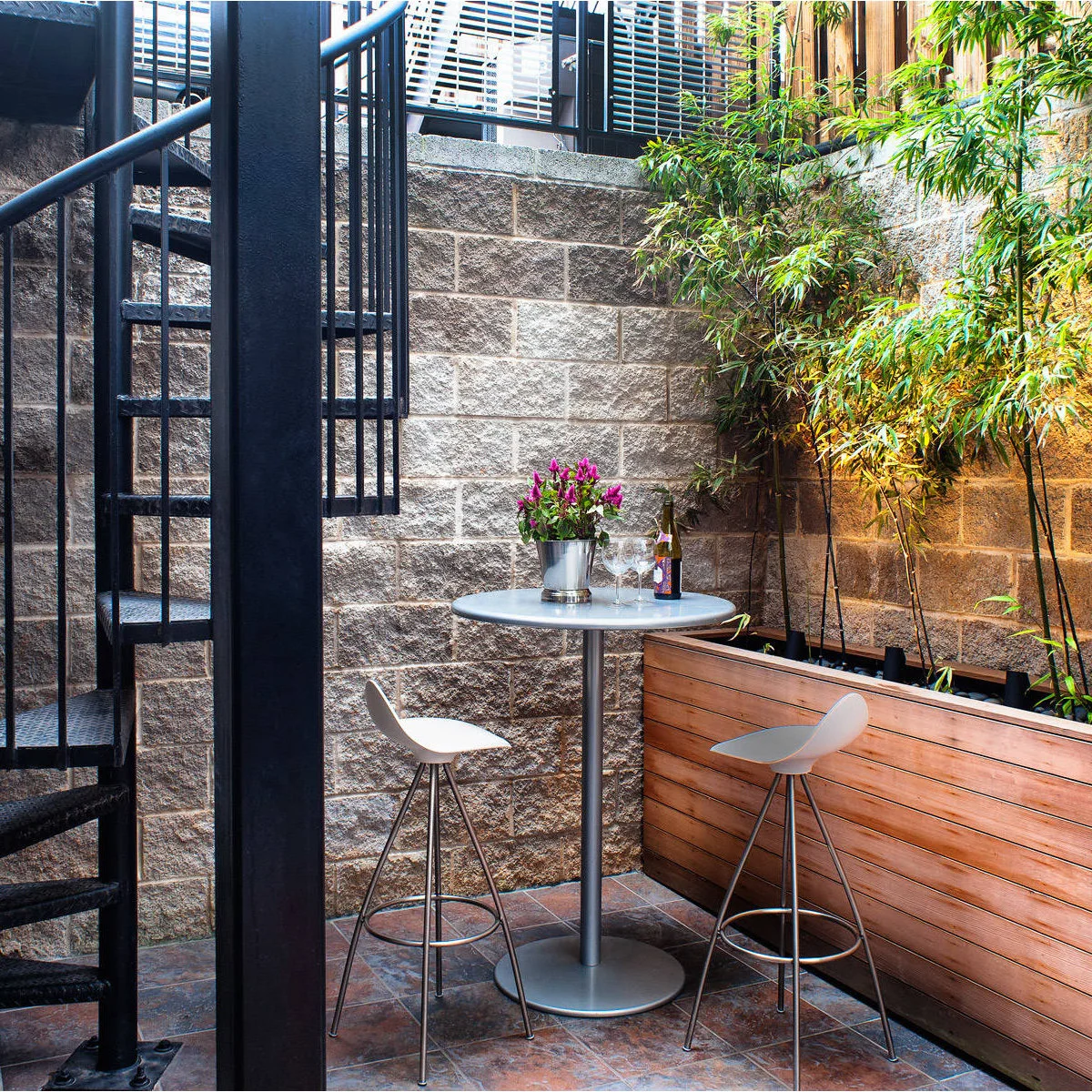 Two pale stools sit at a round high table beside a tall wood planter and masonry walls under an exterior spiral stair.