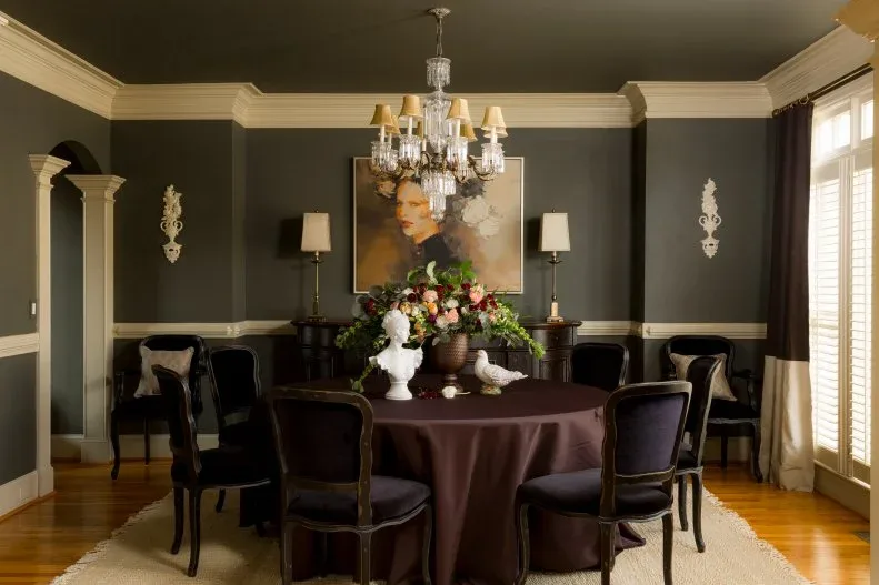 Dark-walled dining room with a cloth-covered round table, dark chairs, pale trim and a shaded chandelier.