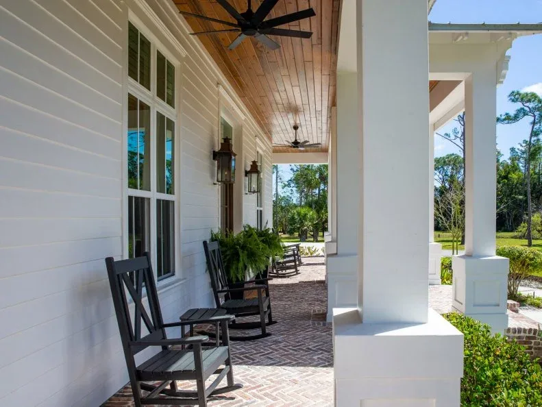 Long white farmhouse porch with wood ceiling, black rocking chairs, fans and brick-pattern floor