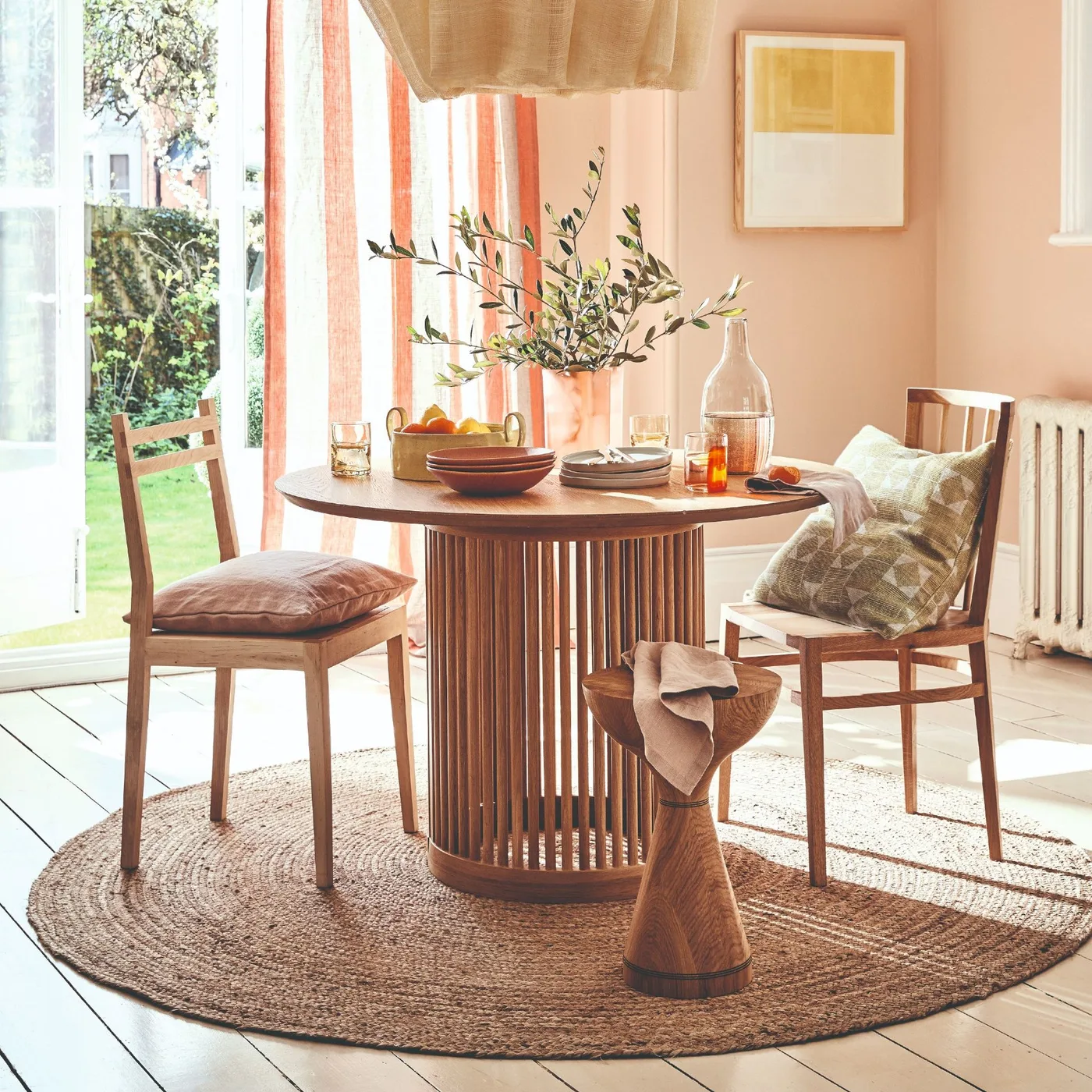 Peach dining corner with a round wooden pedestal table, cushioned wooden chairs, round woven rug and striped curtains.