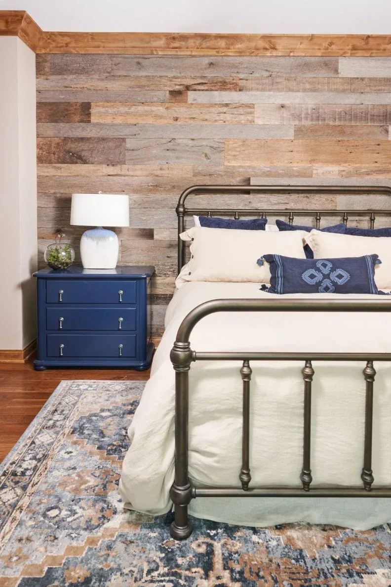 Varied timber wall boards sit behind a dark metal bed beside a blue drawer chest and patterned rug.