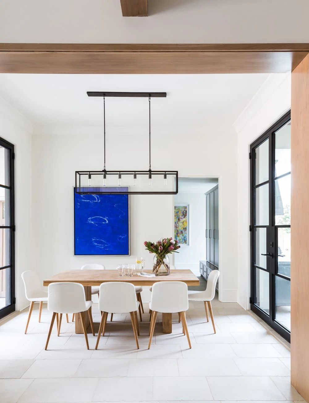 A pale wood dining table and white chairs stand beneath a rectangular black light beside white walls, a blue artwork, wood ceiling beams and black-framed glazed doors