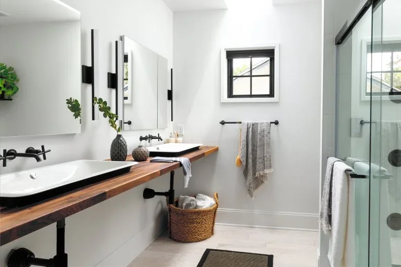 A long wood counter with two shallow basins and dark wall-mounted taps sits beneath tall rectangular mirrors beside a glass shower and white walls