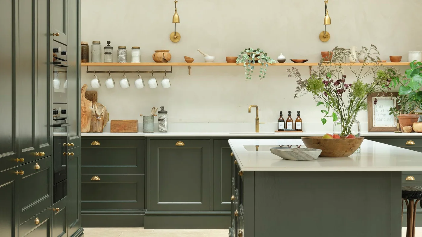 Long wooden shelf with bowls, jars and trailing greenery above a mug rail in a green kitchen.