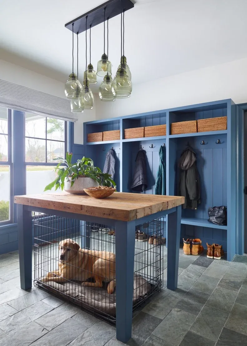 Navy mudroom lockers with woven baskets, coat hooks, a wood-topped central unit with a dog kennel below, windows, and glass pendants.