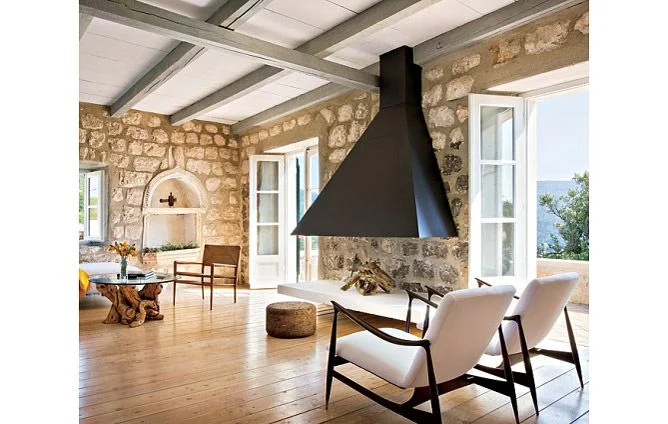 A plain tapered black fireplace hood hangs above an open fire area beside rough stone walls, white-framed windows, pale wood flooring and two light upholstered chairs