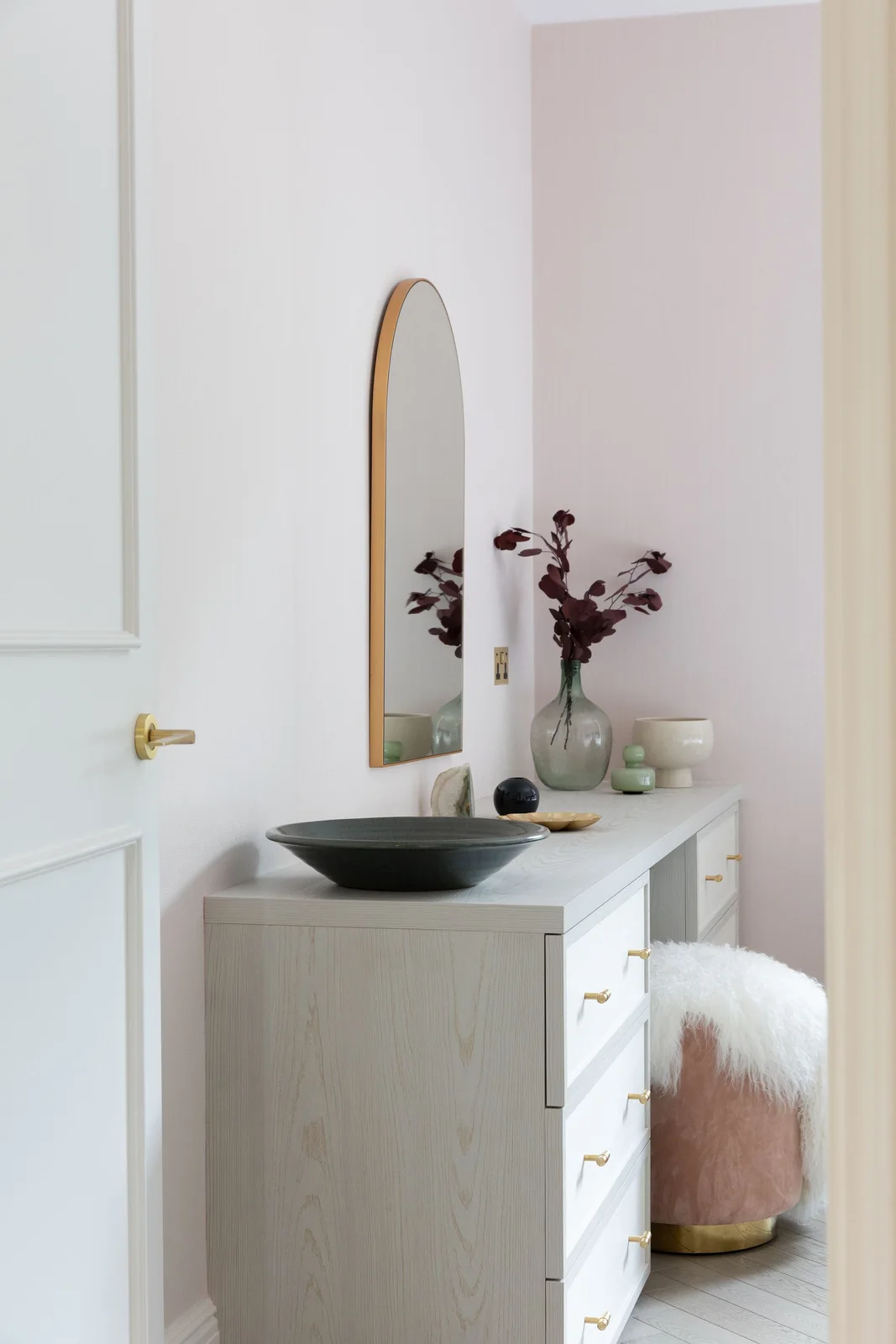 A pale vanity with side drawers and a tall arched wall mirror stands beside a pink upholstered stool