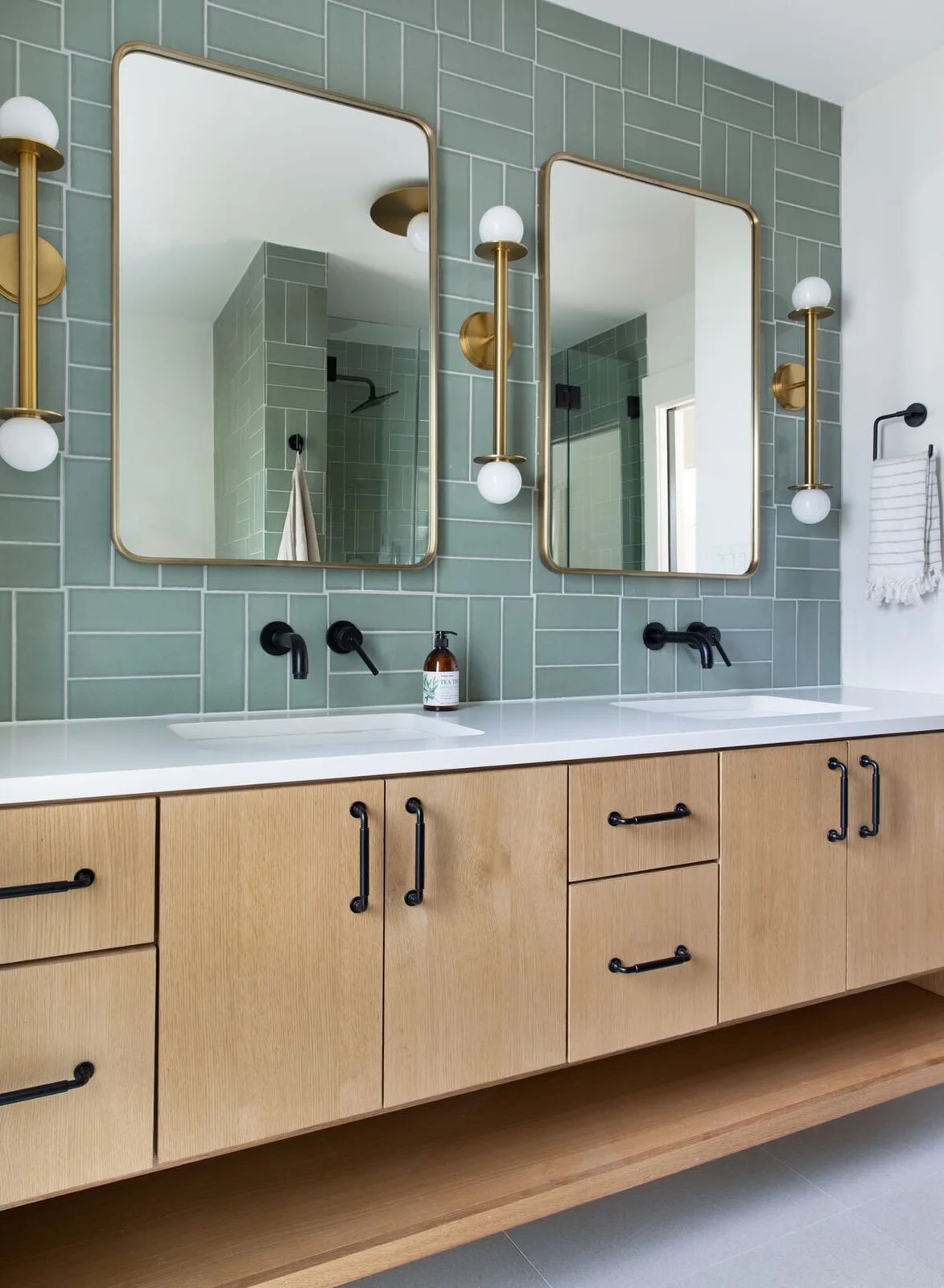 A sage parquet-pattern tile wall backs two gold-framed mirrors and black taps above a long pale wood vanity and white double basin.