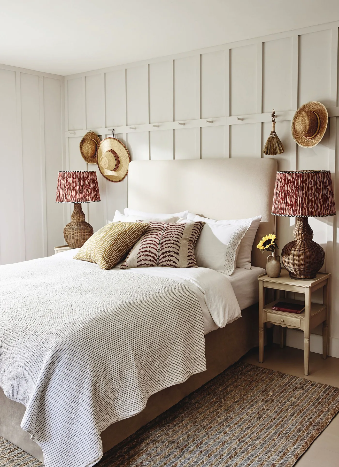 Woven bedside lamps stand against pale wall paneling beside a cream bed, patterned cushions and timber bedside tables.