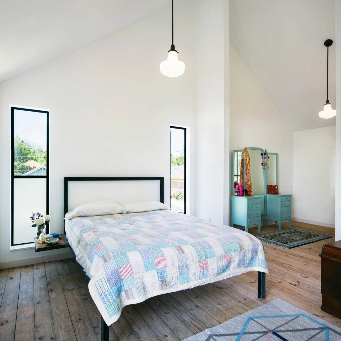 A slim black bed frame stands between narrow dark-framed windows beneath hanging exposed bulbs, with a pastel quilt and turquoise dressing table