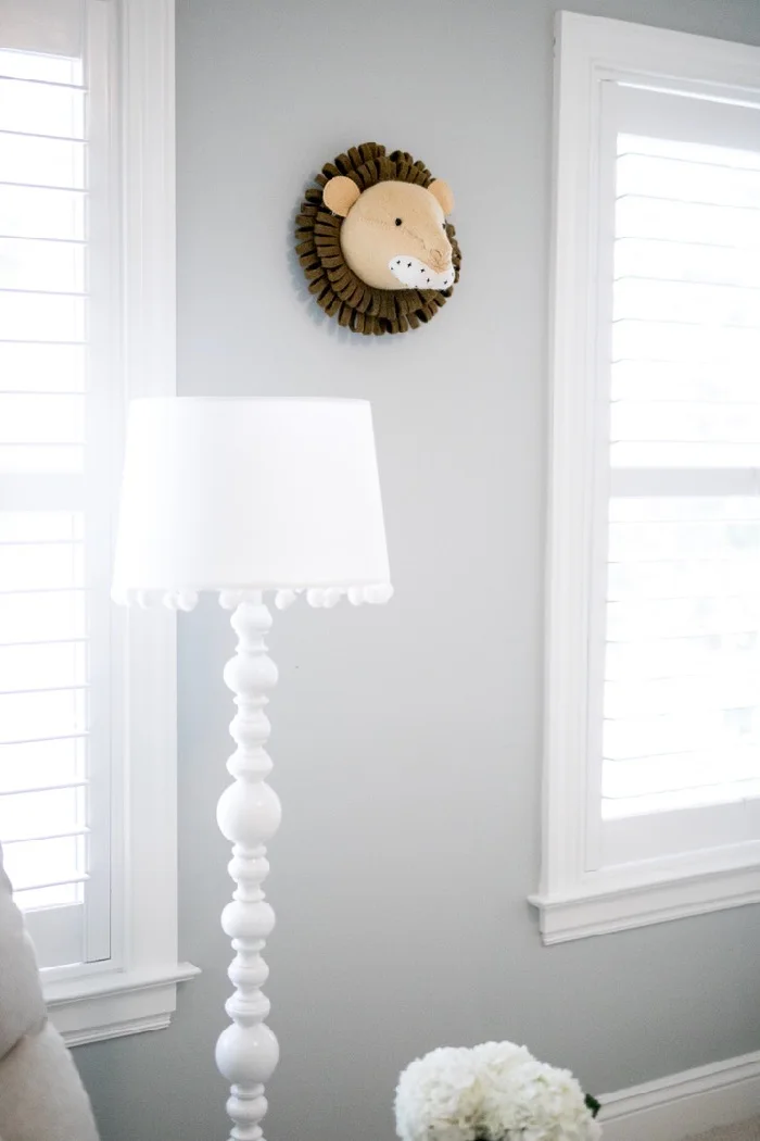 A small soft lion-head wall decoration hangs between shuttered windows above a white floor lamp.