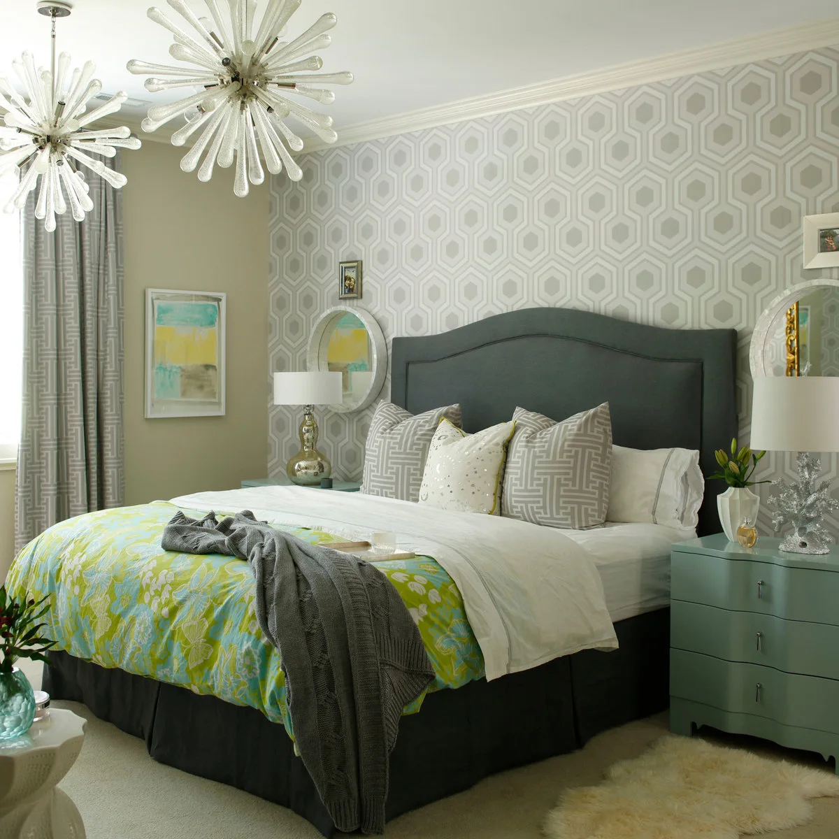 Pale hexagonal wallpaper behind a dark gray curved headboard with patterned gray cushions and turquoise bedside drawers.