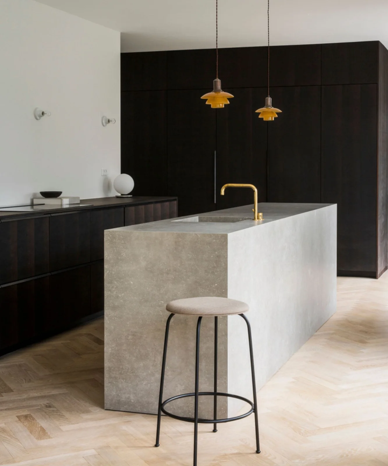 A pale rectangular island with a gold-toned tap stands beside dark cabinet walls, a light stool and two small yellow pendant lights