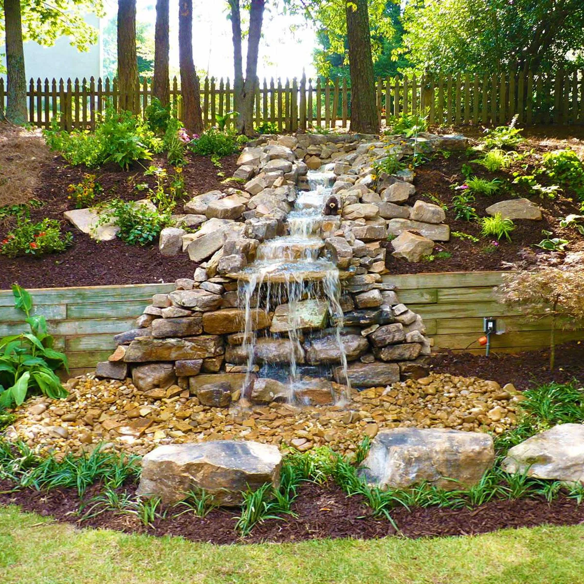 A narrow stepped rock waterfall running down a planted bank, over a wood retaining edge and into a pebble-covered base.