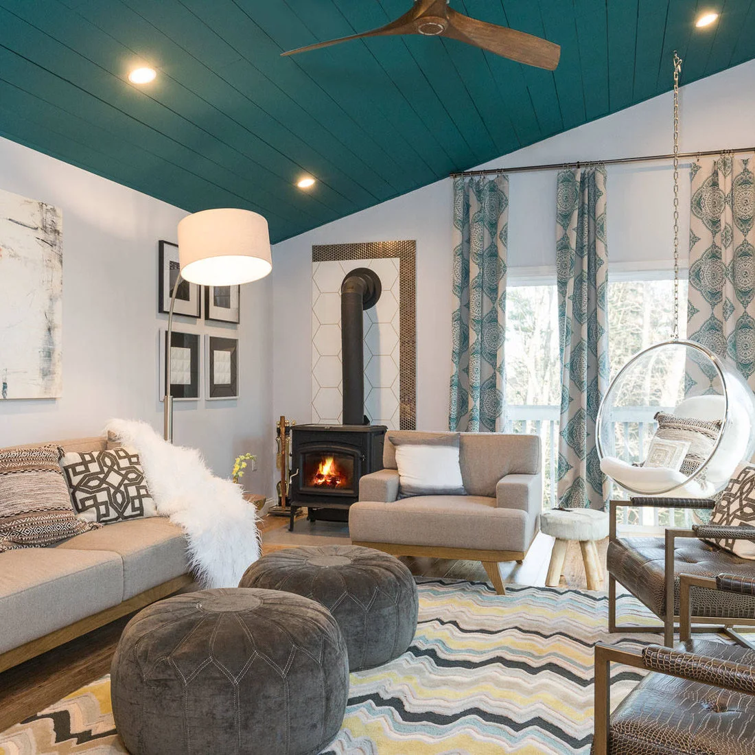 A dark stove stands before a pale tiled corner beneath a teal boarded ceiling, with gray seating, patterned curtains, round pouffes and a clear hanging chair.