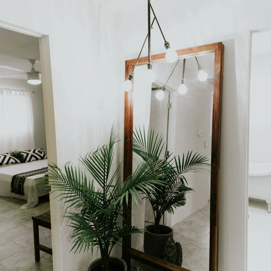 A tall wood-framed mirror reflects a pale hallway and branching ceiling light beside open room doorways and a leafy plant.