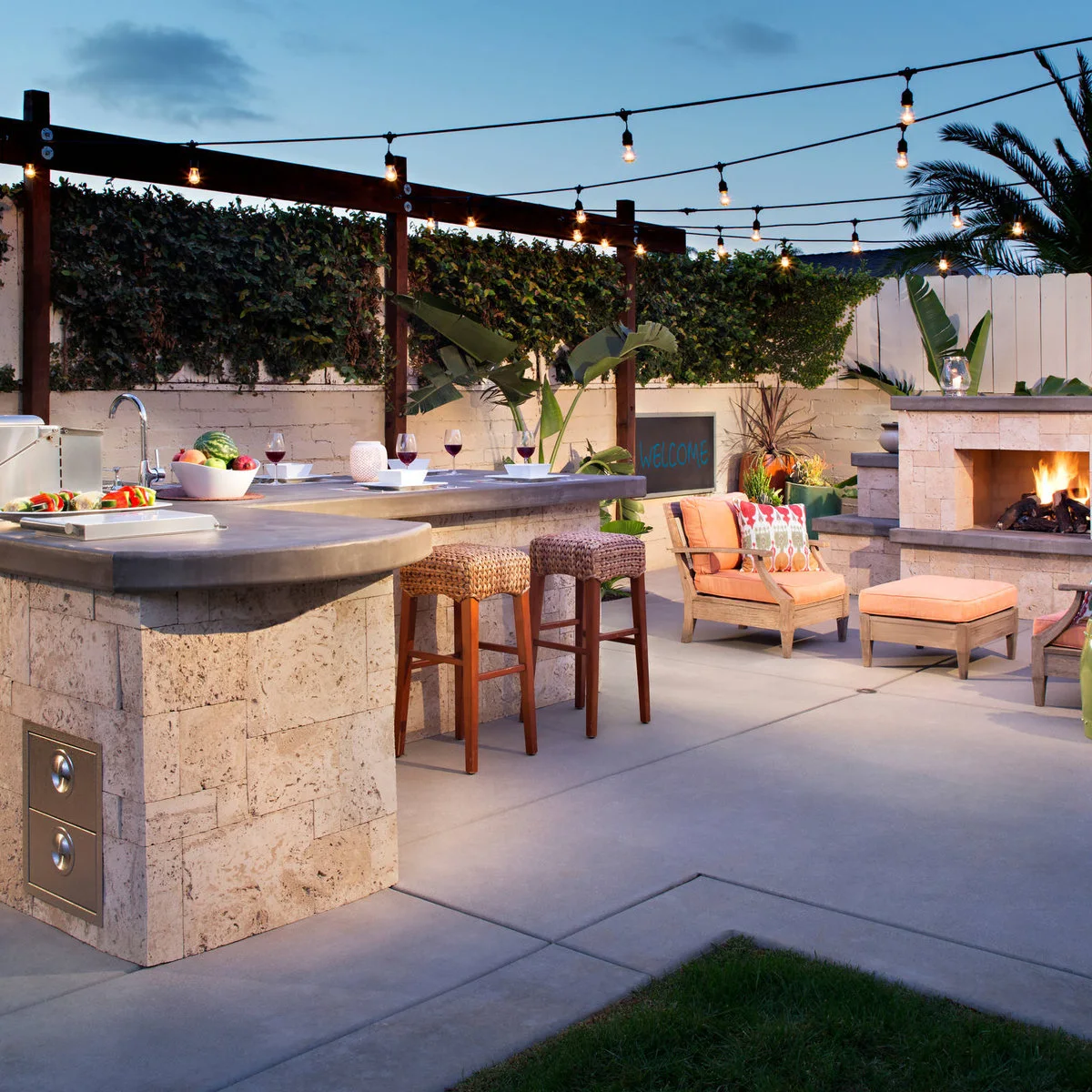 Outdoor kitchen with curved dark serving counter, woven stools, small sink, string lights and nearby fireplace seating