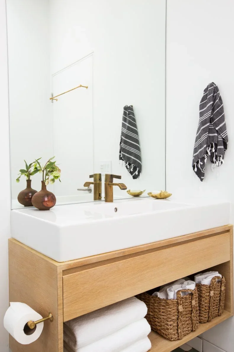 A white basin sits on a pale wood vanity with an open lower shelf holding folded towels and woven baskets beneath a wide mirror