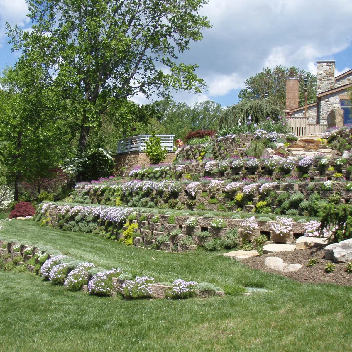 A planted block retaining system covers a sloping garden with trailing flowers, shrubs and nearby stone steps.
